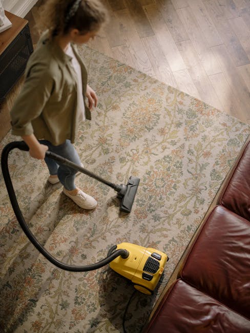 A child using a vacuum cleaner with a yellow body and black hose on an ornate, patterned area rug, part of a domestic setting with soft lighting. The child is standing on the rug, wearing light-colore